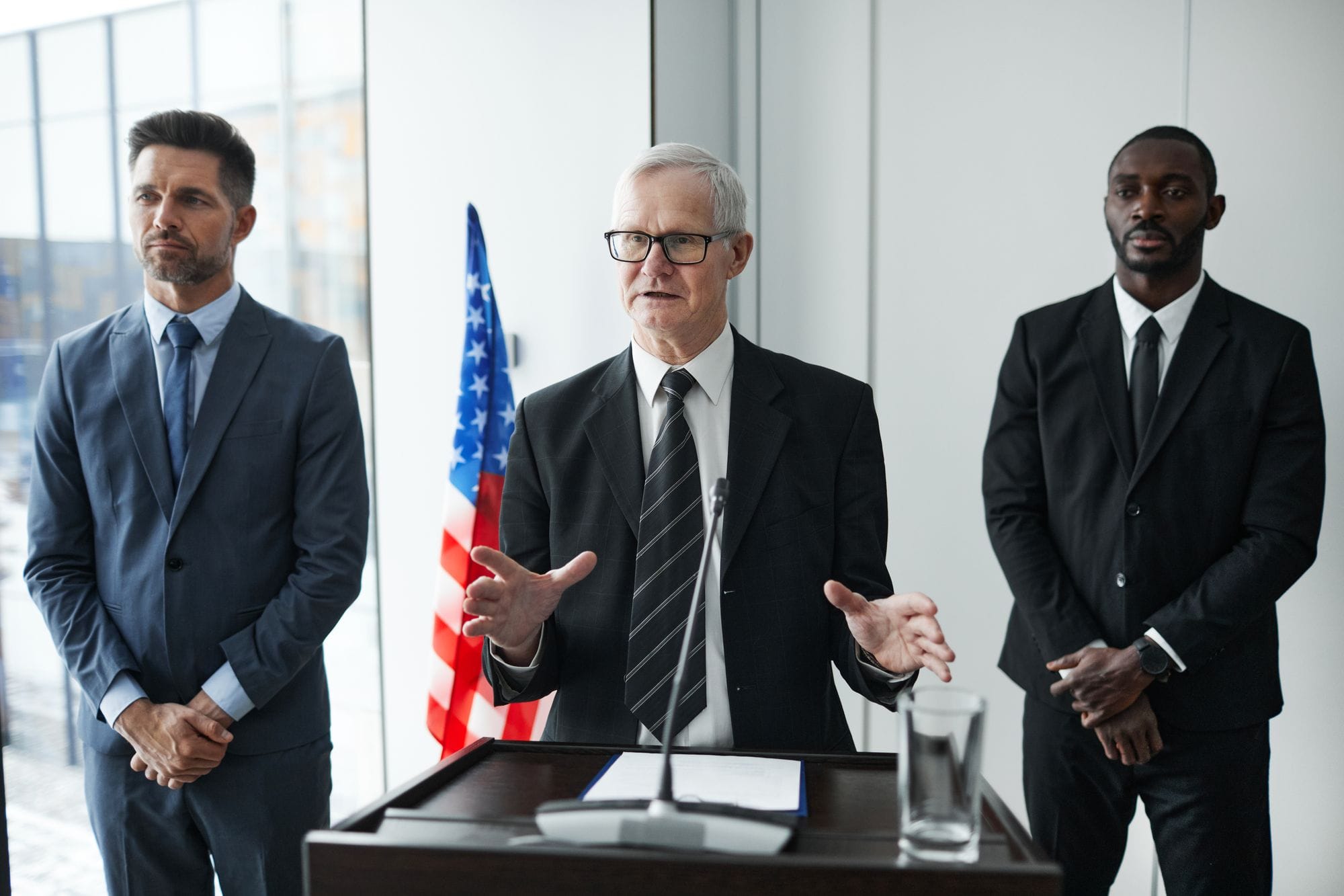 Three business professionals giving a speech with an American flag backdrop.